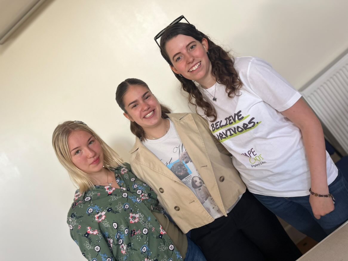 A photo of three smiling caucasian women, the one on the right is wearing a Rape Crisis tee-shirt