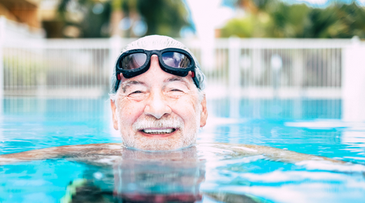 An older man smiles at the camera while swimming in a pool. He has goggles on his head.