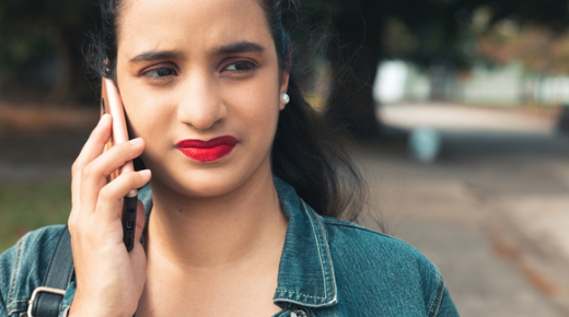 A young woman stands outside. She is talking to someone on her smartphone and is frowning.