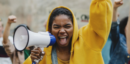 A woman activist shouts into a megaphone, while raising her fist in the air. Other activists can be seen in the background, also raising their fists.