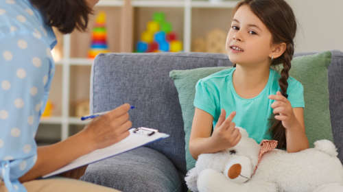 A young girl with pigtails sits on a sofa, holding a stuffed toy and talking to a woman counsellor who is sat on a chair in front of her.