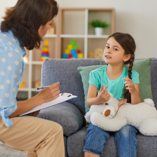 A young girl with pigtails sits on a sofa, holding a stuffed toy and talking to a woman counsellor who is sat on a chair in front of her.