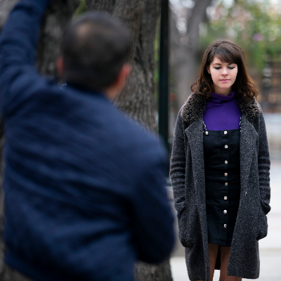 A young woman walks down a street towards us looking nervous as a man stands with his back to us, watching her.