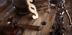 A bronze statue of Lady Justice is in focus. The statue is on a wooden judges desk, out of focus is a wooden gavel and some books.