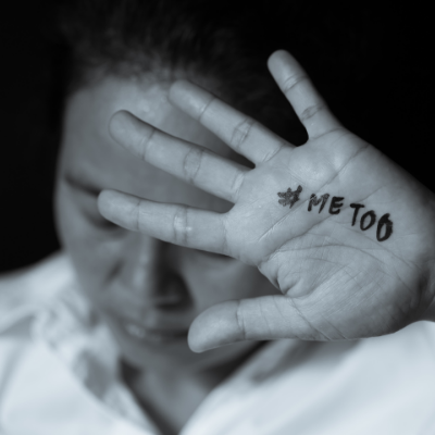 This black and white image shows a woman looking down while holding the palm of one of her hands up to the camera. On her palm is written #METOO.