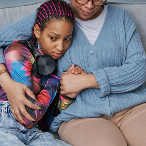 A woman sits on a sofa, tightly hugging a young teenage girl into her.