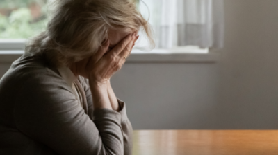 An older woman sits at a table with her head in her hands. Her glasses sit on the table in front of her.