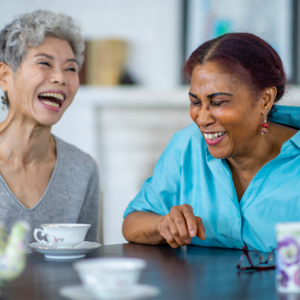 Two older women sit at a table with tea cups on it and are laughing together.