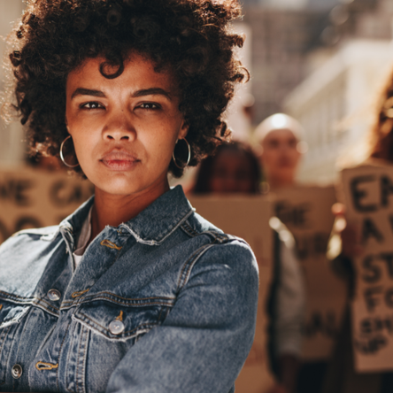 A woman at a women's march stands with her arms crossed looking powerful. Behind her, there is a group of blurred women who are holding up signs.