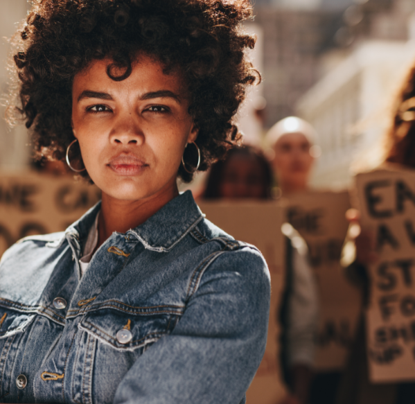 A woman at a women's march stands with her arms crossed looking powerful. Behind her, there is a group of blurred women who are holding up signs.
