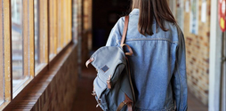 A teenage girl walks down a school corridor. She is wearing a denim jacket and has a backpack slung over one of her shoulders. The photo is taken from the back so we can't see her face.