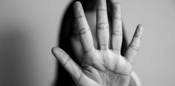 A black and white image of a woman standing opposite a camera.  Her hand is held up in a stop sign, hiding her face.