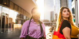 Two young women stand outside in the sunshine. One stands front on to the camera wearing a pink shirt and braids. On her left stands another young women, standing side on facing the camera, holding a rainbow tote bag.