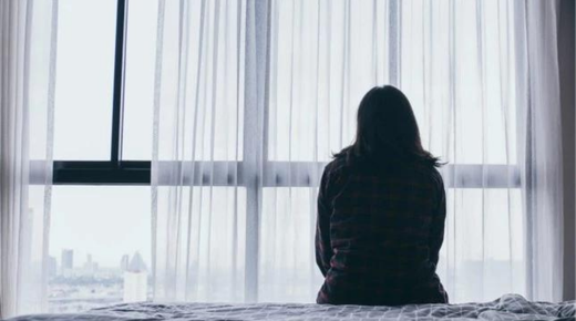 This black and white image shows the back of a woman, sitting on a bed looking out the window.