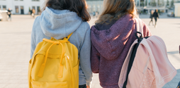 Two young girls from the back standing outside. One has a yellow backpack on, and the other has a light pink spotty backpack on.