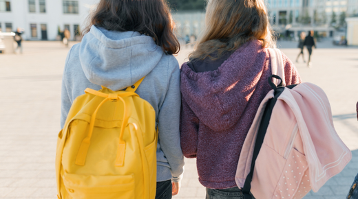 Two young girls from the back standing outside. One has a yellow backpack on, and the other has a light pink spotty backpack on.