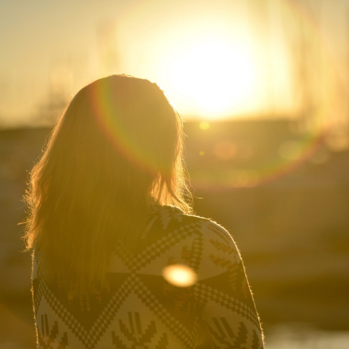 A woman with shoulder-length brown hair stands with her back to us. She is outside and the sun is low in the sky.