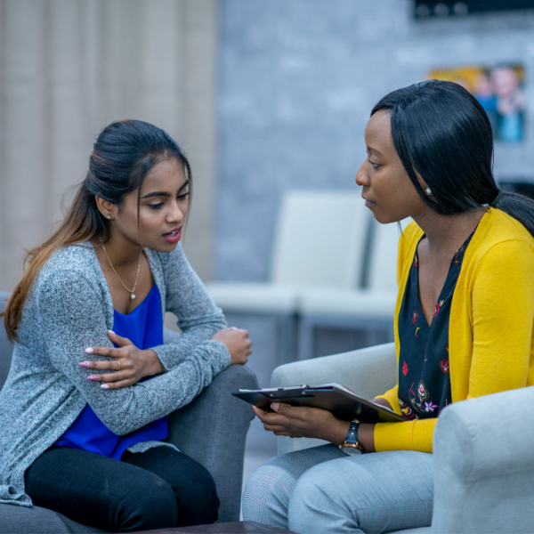 A female counsellor holding a clipboard and her female patient sit on armchairs. The female patient is mid speech and looking at the clipboard and the counsellor is looking at her.
