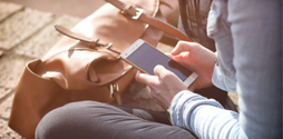 A woman sits cross legged on the floor outside using her smartphone. Her head is not in the shot.