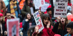 A blurred group of women marching at a protest. Some women are holding up signs, but only one can be read: 'My little black dress doesn't mean yes'.