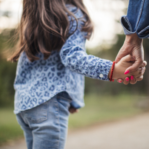 A young girl with long brown hair holds the hand of a woman who is off camera. Only the back of the girl can be seen.