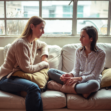 A young girl sits cross-legged on a sofa opposite her mother. The girl is talking while the woman looks serious.
