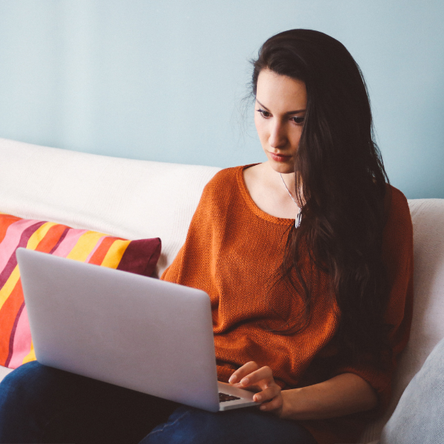A young woman sits on a sofa and looks at a laptop.