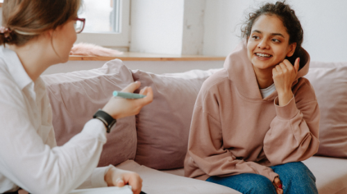 A young woman wearing a beige hoodie and blue jeans talks to a young female counsellor. Both are sitting on a sofa.