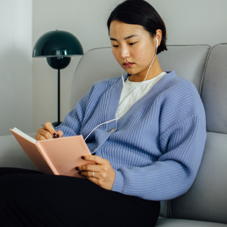 A young woman sits on a sofa, writing in a journal with her headphones in.