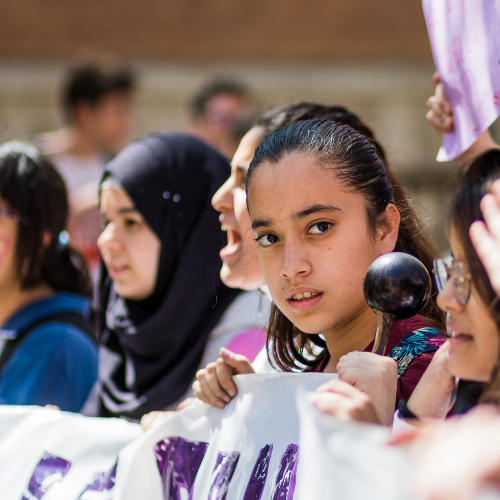 A group of young women and girls of different ethnicities are pictured at a protest holding a giant banner and posters.